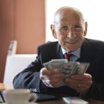 Investing For Retirement - Confident senior businessman holding money in hands while sitting at table near laptop