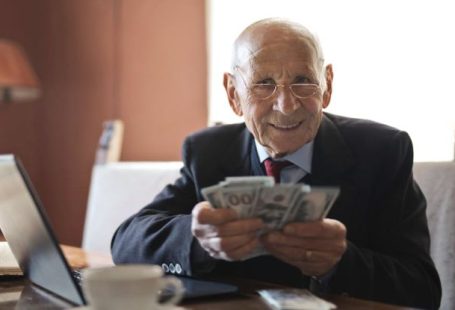 Investing For Retirement - Confident senior businessman holding money in hands while sitting at table near laptop